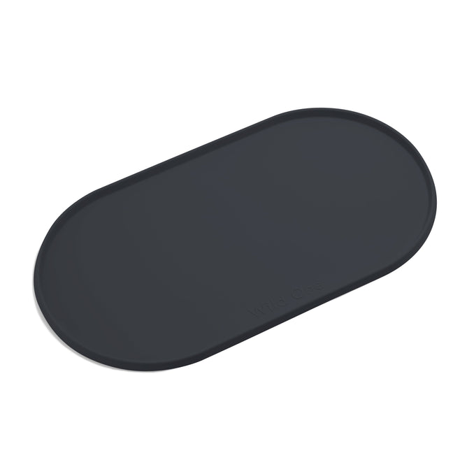 The Wild One Silicone Pet Placemat is a dark gray, oval-shaped mat with a subtle embossed logo and a raised lip for spill prevention, making it easy to clean.