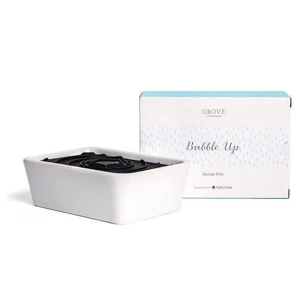 A white rectangular sponge dish with a black insert, displayed beside its packaging, showcases a simple design with light blue accents. The box reads Grove Co. Bubble Up Dish Soap Dispenser Sponge Dish. Made from durable ceramic, it adds sleekness to any kitchen.