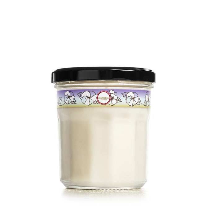 Mrs. Meyers Soy Candle comes in a glass jar filled with white cream and topped with a black lid. The label showcases floral illustrations on a light purple background with a central red circle, evoking a garden-fresh fragrance.