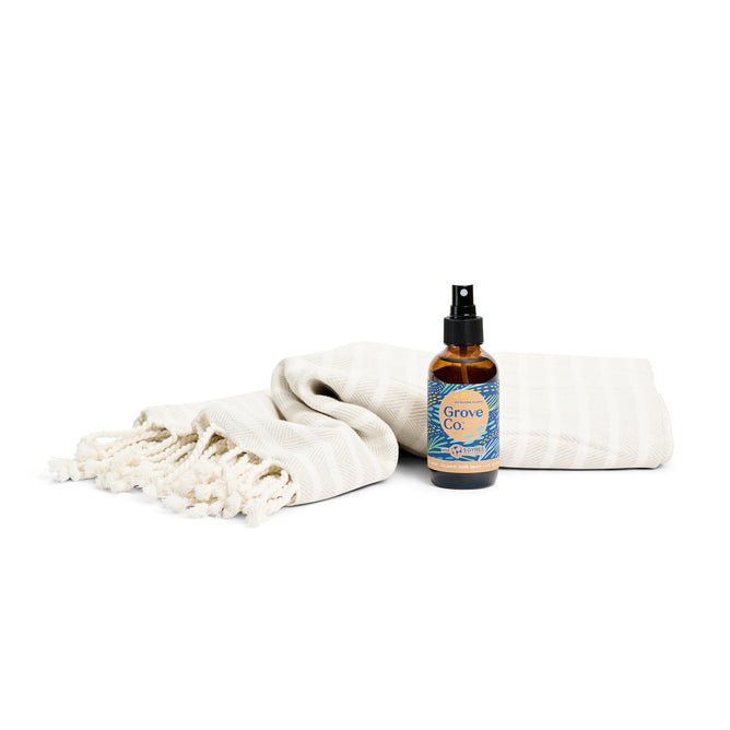 A white fringed towel and a small amber spray bottle labeled Grove Co. from the Bathroom Refresh Set - Spring Collection by Grove Collaborative, featuring the limited edition scent Atlantic Dune Grass, are displayed against a plain white background.