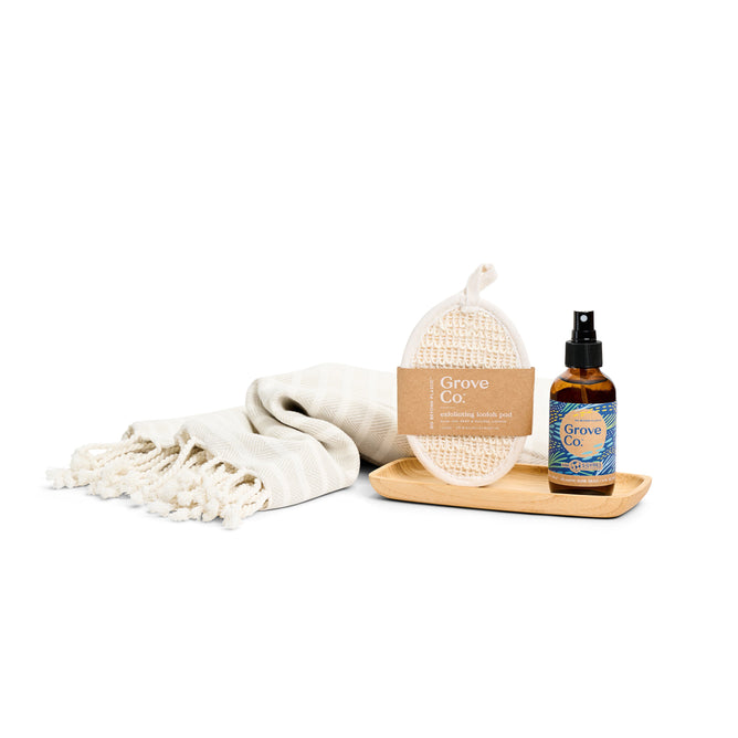 The Grove Collaborative Refresh & Relax Set from the Spring Collection showcases a neatly folded beige towel with fringed edges, a textured loofah with a label, an Atlantic Dune Grass spray bottle, and a light wooden tray, all beautifully arranged on a white background.