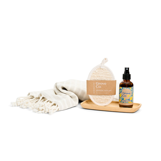 The Refresh & Relax Set - Spring Collection by Grove Collaborative includes a white woven towel with fringed edges, a textured bath pad in a cardboard sleeve, a wooden tray, and a natural fragrance spray. These items are neatly arranged on a white background.