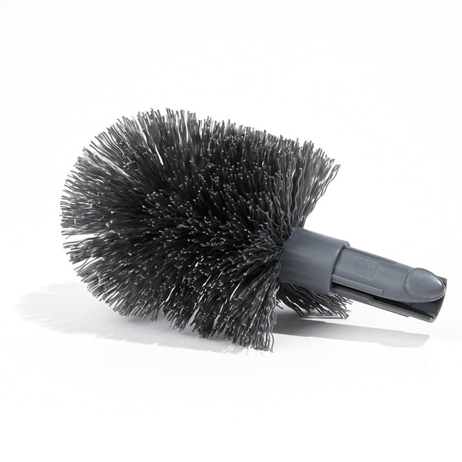 A round, dense black Grove Co. Toilet Brush Head Refill rests on a white surface, designed to be replaceable for cleaning purposes. It has an attachment slot to fit handles or devices, aiding in reducing plastic waste. Shadows cast left suggest lighting from the right side.