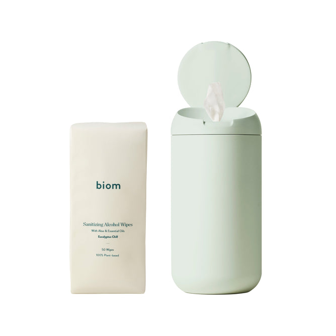 A pastel green cylindrical container with a lid open reveals CDC-compliant sanitizing wipes. Beside it is a matching Biom pack labeled Hand Sanitizing Wipes Starter Kit, featuring Sanitizing Alcohol Wipes with Aloe, Coconut Oil, and Eucalyptus Oil—50 plant-based wipes for every clean need.