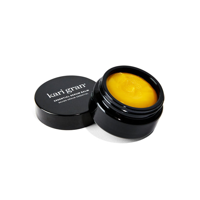 A black container of Kari Grans Essential Serum Balm is open, revealing the ultra-hydrated yellow balm. The lid, featuring the brands logo, rests beside it, elegantly displayed against a pristine white background.