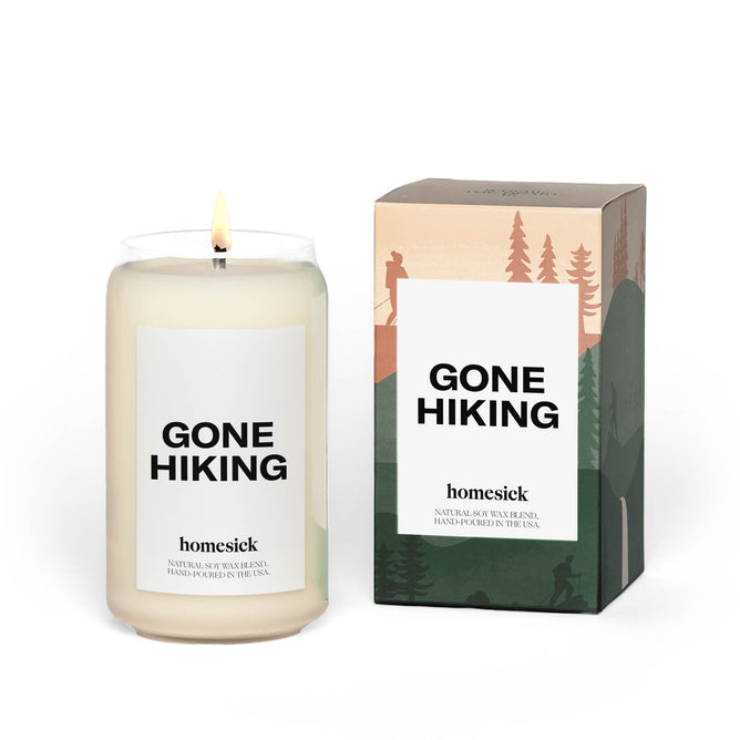 The Homesick Gone Hiking candle, crafted from natural soy wax, is lit beside its box. The white jar has a simple label, and the box features outdoor-themed illustrations with mountains and hikers. Enjoy sandalwood jasmine notes in this cruelty-free creation.