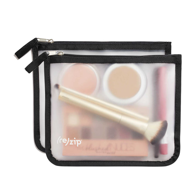 Two transparent (re)zip Zippered Medium Bags with black edges and zippers display visible makeup items like a round compact, tube, and brush. The travel bags are stacked slightly offset in their sleek translucent design, with one partially covering the other.