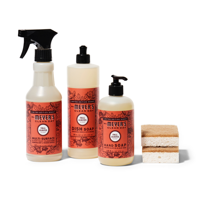 The Grove Collaborative Happy Harvest Essentials Set - MMCD features three Mrs. Meyers Clean Day products in the Fall Leaves scent: a multi-surface spray, dish soap, and hand soap. Two rectangular sponges accompany them, all set against a white background.