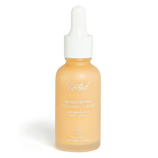 A frosted glass bottle with a white dropper cap features Rooted Beauty Brightening Vitamin C Serum, showcasing its antioxidant-rich essence. Infused with kakadu plum and R7 complex, this lightweight serum is powered by seven antioxidant-rich roots and casts a pale orange hue.