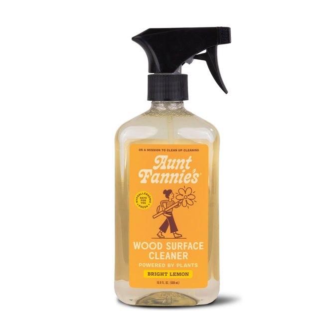 Aunt Fannies Wood Surface Cleaner comes in a spray bottle with bright lemon scent and essential oils. The label, featuring a person and flower, emphasizes plant-powered surfactants, while the bottle contains clear liquid.