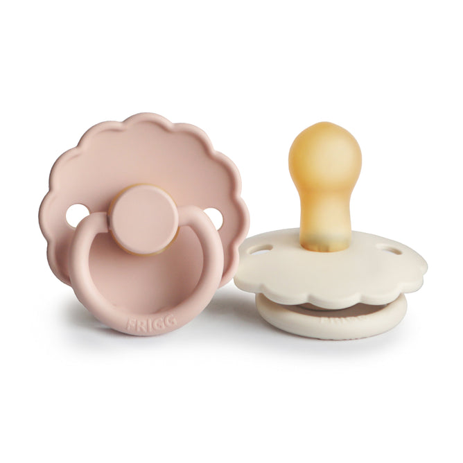 Two mushie FRIGG Daisy Natural Rubber Pacifiers (BPA-Free, 0-6 months)—one pink and flat, the other white and upright showing its tan nipple—boast charming Danish design with scalloped edges.