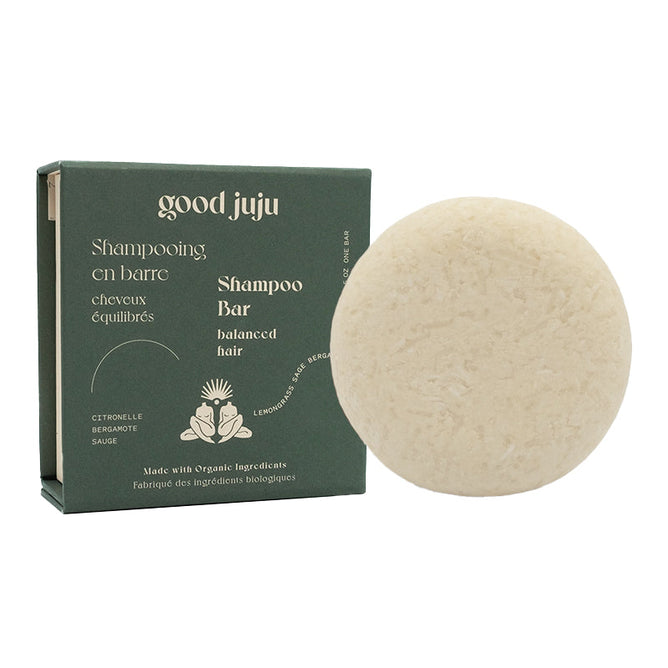 A round, off-white shampoo bar sits beside an open dark green box labeled in English and French. The box showcases the Normal/Balanced Shampoo Bar by Good Juju Body & Home, highlighting its all-natural citronella, bergamot, sage, and coconut oil blend for perfectly balanced hair.