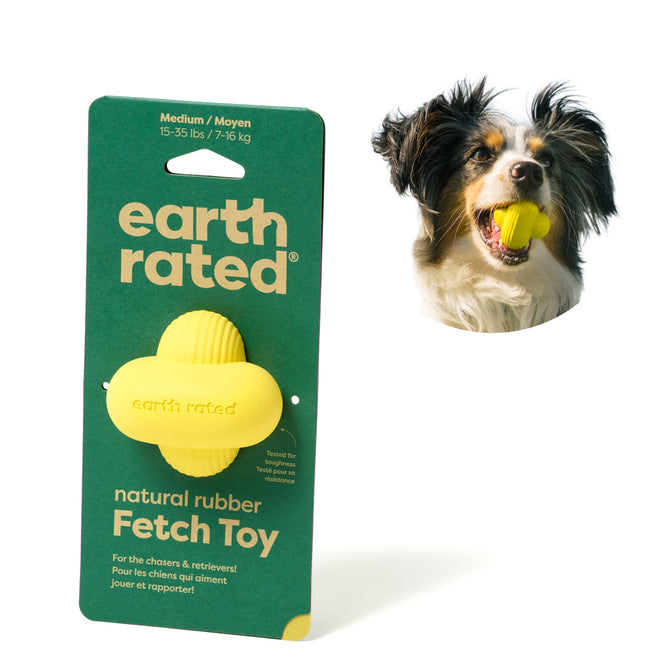 A dog with fluffy ears holds an Earth Rated Rubber Fetch Chew Dog Toy (Medium) in its mouth. Next to it is the packaging for this medium-sized toy, ideal for dogs weighing 15-35 lbs (7-16 kg).