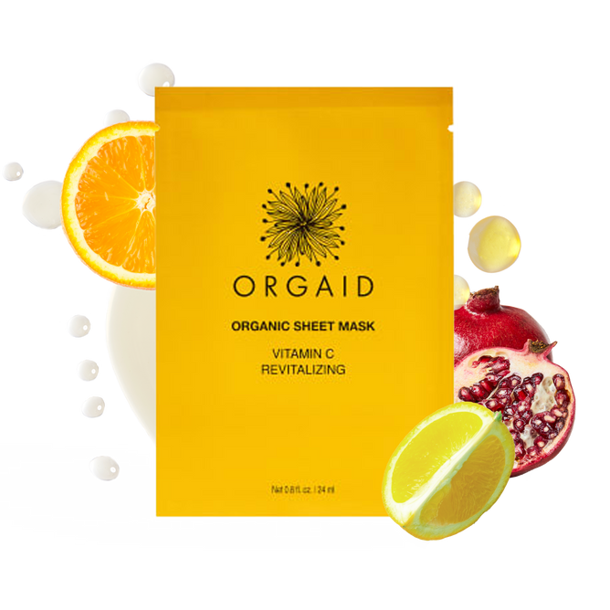 The image features an ORGAID Vitamin C & Revitalizing Organic Sheet Mask in a vibrant yellow package, surrounded by orange, lemon, and pomegranate slices. The mask uses Ecoderma fabric technology for optimal absorption amidst liquid splashes.