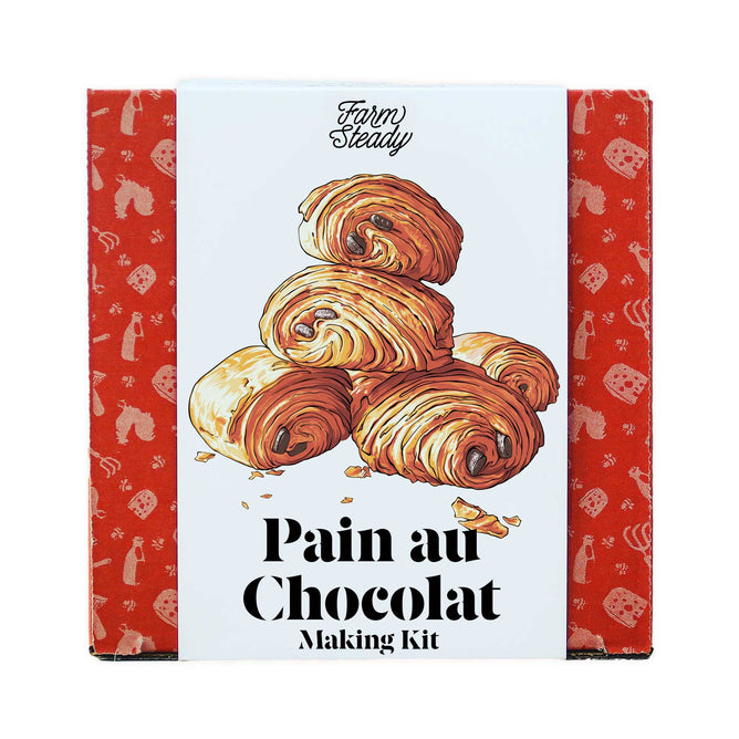 FarmSteadys Pain Au Chocolat Making Kit box showcases an illustration of stacked French pastries, with a backdrop of croissants, coffee cups, and mugs. It includes premium Dark Chocolate Batons for an authentic touch.