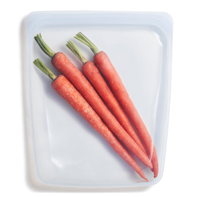 Four fresh carrots with green tops are neatly stored in a Stasher Reusable Silicone Half Gallon Bag on a light surface, highlighting an eco-friendly storage solution.