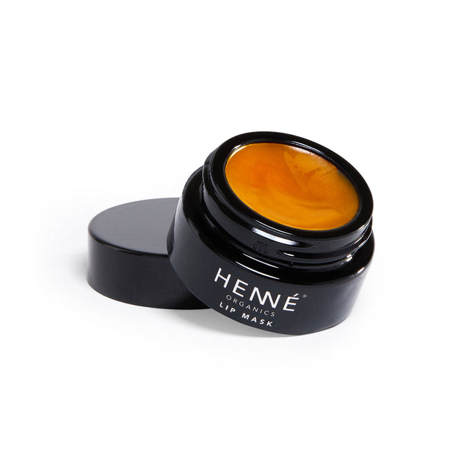 A Henné Organics Lip Mask jar, with its black exterior, reveals a rich golden-orange balm inside made from organic ingredients. The lid is placed beside the jar, showcasing its glossy texture against a simple white background.