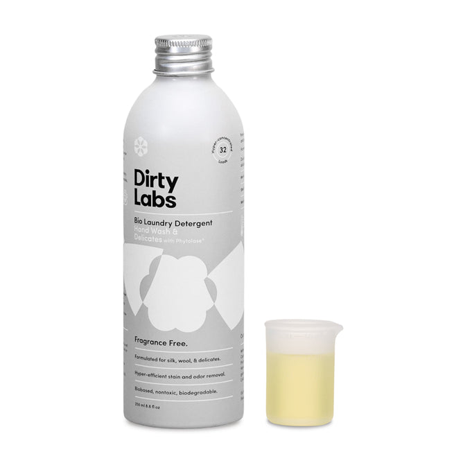 A silver bottle of Dirty Labs Bio Laundry Detergent with Phytolase® for Hand Wash & Delicates, part of our eco-friendly laundry supplies, stands next to a small measuring cup with light yellow liquid. Its fragrance-free and ideal for those seeking natural laundry detergents.