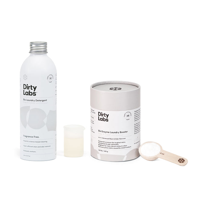 A Dirty Labs scene features the Bio Laundry Detergent with Phytolase® in a white bottle with a silver cap beside a cylindrical Bio Enzyme Laundry Booster, highlighting clean science. A small measuring cup and wooden scoop with enzyme-rich powder complete the set.