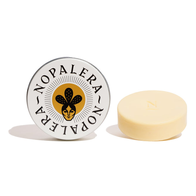 A tin of NOPALERA Moisturizing Botanical Bar features a yellow lid with a stylized face and plant logo. The light yellow bar sits on a white surface, ideal for sustainable storage.