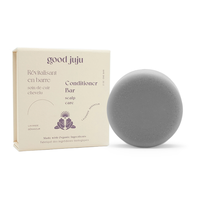 A round gray Scalp Care Conditioner Bar from Good Juju Body & Home, scented with Lavender Geranium, sits next to a beige box featuring a lavender illustration. It uses organic botanicals to relieve dandruff.