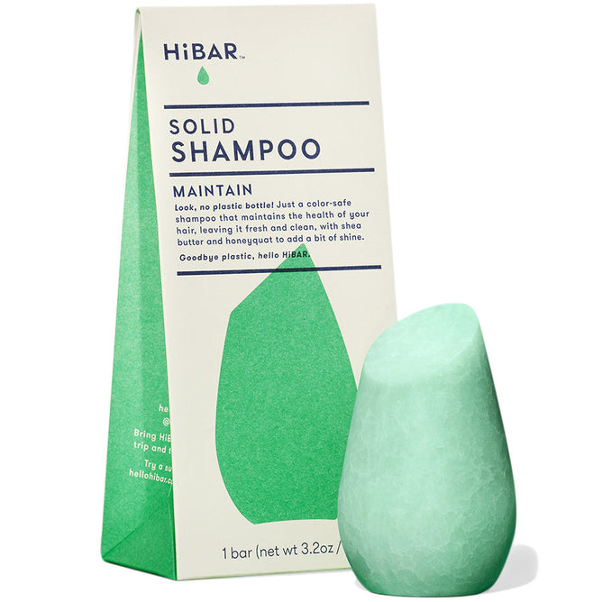 A green teardrop-shaped HiBAR Maintain Solid Shampoo bar is placed in front of its triangular packaging, emphasizing it as a plastic-free shampoo that supports natural hair care and keeps color-treated hair healthy.