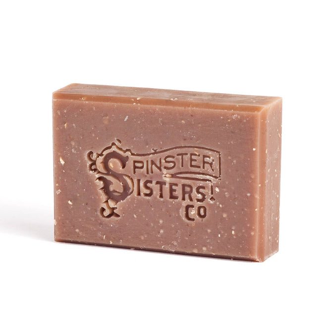 A rectangular Spinster Sisters Co. Bar Soap features a textured, exfoliating surface with an embossed logo. This vegan soap has a natural, rustic look and a brownish color with tiny specks.