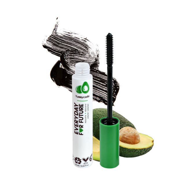 A tube of Everyday For Futures Volumizing Avocado Mascara, with sustainable packaging and a green cap, sits beside a halved avocado. The brush extends outside, and black mascara streaks are smeared in the background.